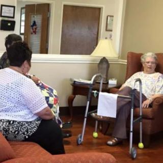 An elderly woman seated in a nursing home, engaged in conversation with visitors.