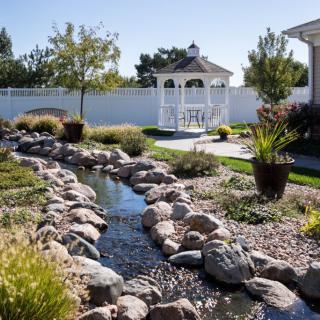 A beautiful garden with a stream flowing through rocks, surrounded by lush greenery and featuring a gazebo in the background.