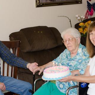A family celebrates a birthday with an elderly woman holding a cake indoors.