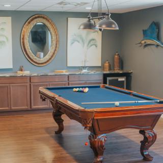A stylish billiard room featuring a pool table with blue felt, hardwood floors, and elegant decor including a mirror and artwork.