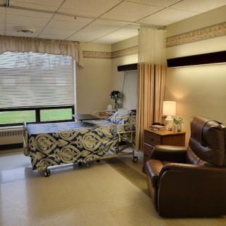 A comfortable hospital patient room featuring a bed, a chair, and natural light from the window.