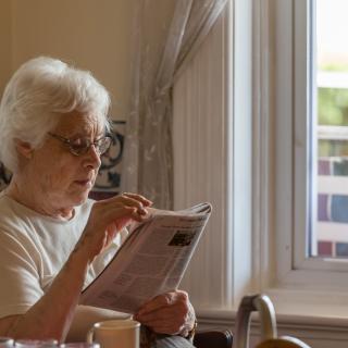An elderly woman reads a newspaper by the window in daylight.