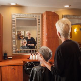 A hairdresser works on a senior woman's hairstyle in front of a mirror at a salon.