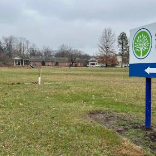A view of an open grassy area with a directional sign pointing left, set against a backdrop of cloudy skies and trees.