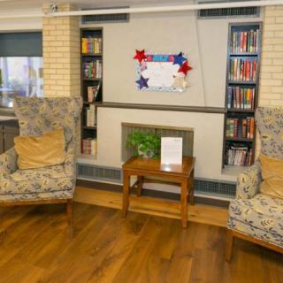 A cozy living room with two patterned armchairs flanking a small wooden table, surrounded by bookshelves and centered around a decorative fireplace.