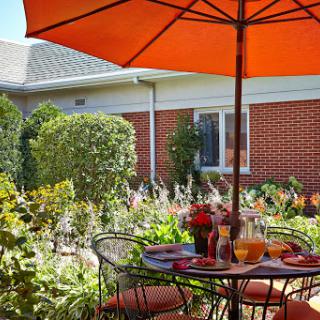 A charming outdoor dining space with a vibrant orange umbrella, surrounded by lush greenery and blooming flowers.