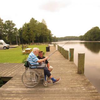 An elderly man in a wheelchair enjoys a moment by the lake, sharing laughter and fishing tips with a friend.