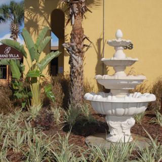 A decorative white fountain in a landscaped garden setting with tropical plants and a building in the background.