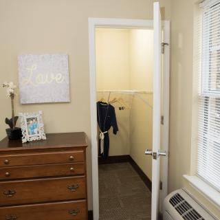 A cozy bedroom featuring a small closet with clothes hanging inside, a wooden dresser, and a window with blinds.