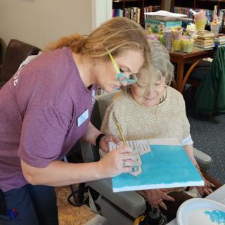 A caregiver assists an elderly person with a painting activity, promoting creativity and interaction.