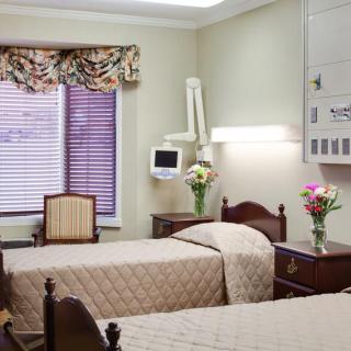 A hospital room with two neatly made beds, a medical monitor, and floral arrangements on the bedside tables.
