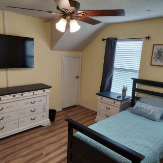 A cozy bedroom with a bed, dresser, wall-mounted TV, ceiling fan, and window with curtains.