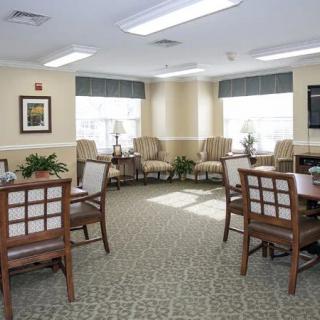 A well-lit room with arranged seating and tables, featuring carpeted flooring and large windows.