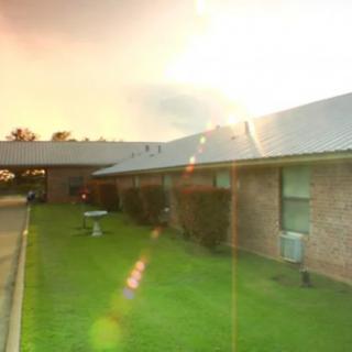 A brick building with a well-maintained lawn is illuminated by the setting sun, creating a warm glow and sun flare effect.