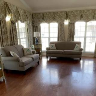 A spacious living room with elegant furniture, featuring a sofa and an armchair on a hardwood floor, adorned with large windows and decorative curtains.