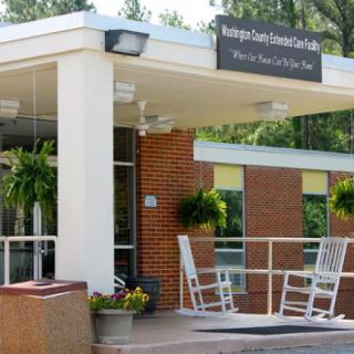 The entrance of a nursing home features rocking chairs and greenery.