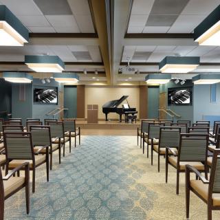 A small concert hall featuring a grand piano on stage with rows of chairs for an audience.