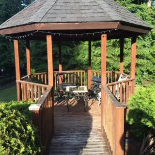 A wooden gazebo in a garden setting, illuminated by sunlight.