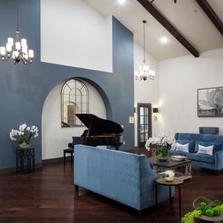 Stylish living room featuring a grand piano and blue furniture, highlighted by elegant chandeliers and a beamed ceiling.