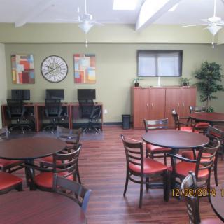A communal space with dining tables and chairs, computers, and a large wall clock.