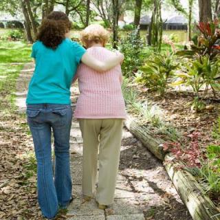 A young woman supports an elderly person while walking in a garden.