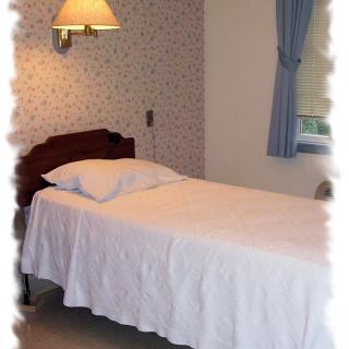 A simple bedroom interior featuring a single bed with white bedding, a wall-mounted lamp, and blue curtains.