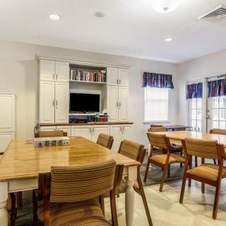 A well-lit dining area with wooden tables and chairs, featuring a television and large windows with curtains.