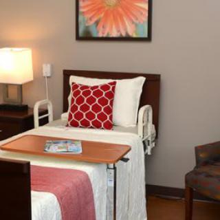 A neatly arranged hospital room featuring a patient bed with a red pillow, a bedside table, and a chair.