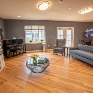 A cozy living room with a wooden floor, featuring a sofa, armchair, coffee table, and piano.