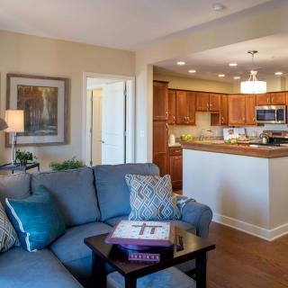 A cozy living room with a view into a modern kitchen, featuring wooden floors and comfortable furniture.