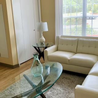 A modern living room with a white sofa, glass coffee table, and a large window allowing natural light to fill the space.