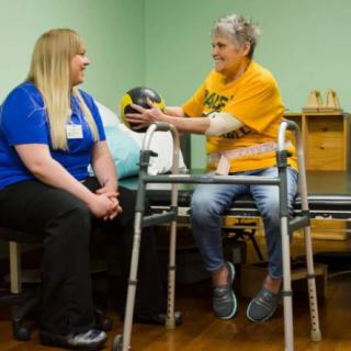 An elderly woman engages in physical therapy with a caregiver assisting her, using a walker and an exercise ball.