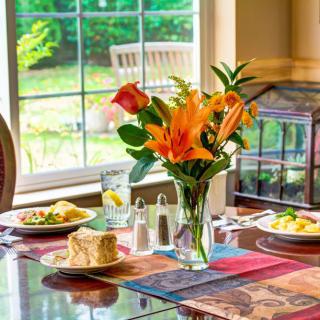 A beautifully set dining table with vibrant flowers and a meal, illuminated by natural daylight from the window.