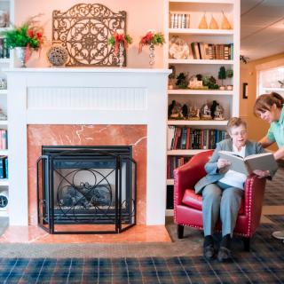 An elderly person reads with the assistance of a caregiver in a cozy living room setting featuring a fireplace and bookshelves.