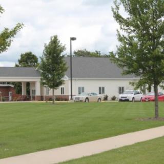 A school building with a grassy lawn, cars parked in front, and trees lining the pathway.