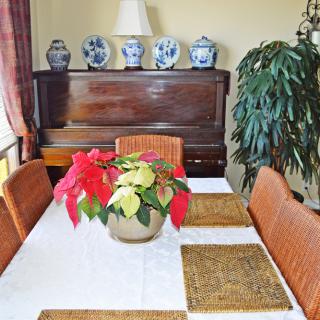A dining room with a table set for four, featuring a poinsettia centerpiece and a piano in the background adorned with decorative vases.