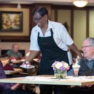 A waitress serves dessert to two senior customers in a restaurant setting.