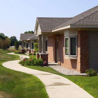 A row of single-story suburban houses with a winding pathway and green lawns under a clear sky.
