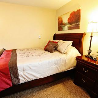 A cozy bedroom with a neatly made bed, warm lighting from a lamp on the nightstand, and a landscape painting on the wall.