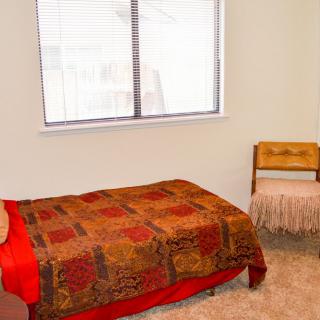 A simple bedroom with a single bed, patterned quilt, and a wooden chair near the window.