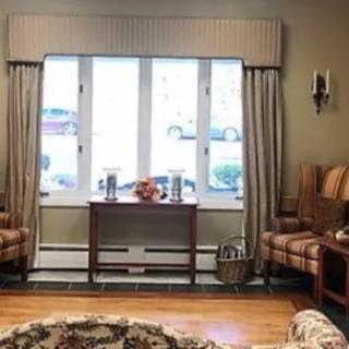 A cozy living room with a window, curtains, chairs, and a table on a wooden floor.