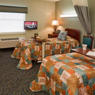 A hospital room featuring two beds adorned with colorful quilted bedspreads, a TV on the wall, and a window with blinds.
