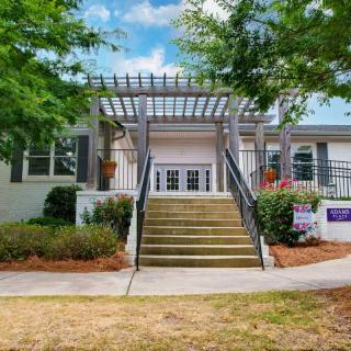 A picturesque building entrance with stairs and a pergola surrounded by greenery.