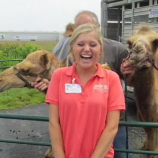A woman smiling while standing in front of two camels at a farm.