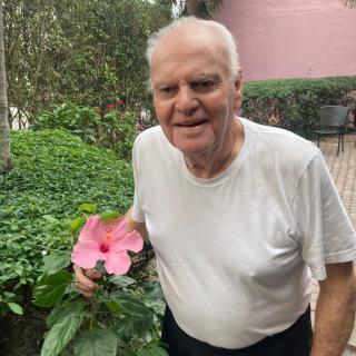 An elderly man smiling while holding a pink flower in a garden.