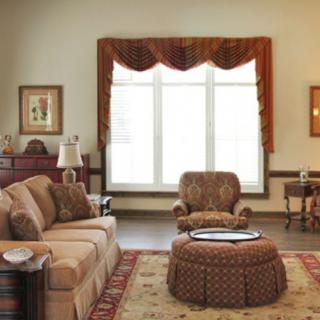 A cozy living room with a sofa, armchair, and ottoman arranged around a central table in front of a large window with drapes.