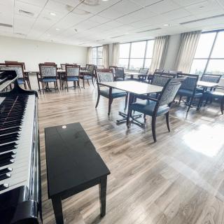 A bright room with a piano, tables, and chairs arranged on a hardwood floor.