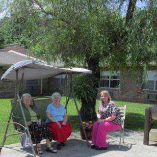 Three individuals sitting on a swing and chair outdoors, enjoying a sunny day under a tree.