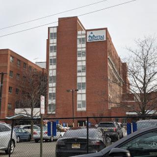 A brick building with multiple stories, possibly a residential or hospital facility, surrounded by parked cars.