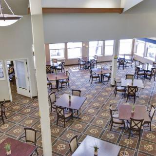 A spacious dining area with multiple tables and chairs, featuring large windows and carpeted flooring.
